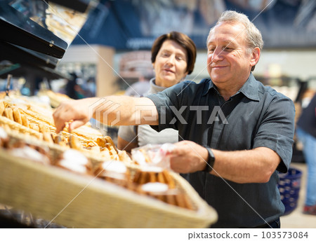 Elderly married couple chooses hot fresh buns and bread at supermarket showcase Elderly married couple chooses hot fresh buns and bread at supermarket showcase 103573084