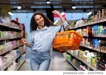 Portrait of a happy woman shopper in a supermarket, a Hispanic woman with a basket of goods smiles with pleasure and dances among the shelves with goods, a satisfied shop customer. Portrait of a happy woman shopper in a supermarket, a Hispanic woman with a basket of goods smiles with pleasure and dances among the shelves with goods, a satisfied shop customer. 103574442