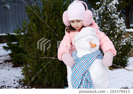 Charming cute little child girl puts on a blue shawl on a snowman, during active winter leisure games in the snowy backyard in the fresh air. Happy childhood and healthy lifestyle. Christmas time 103576913