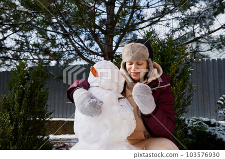 Happy cheerful positive woman making a snowman in the snow covered backyard, putting a carrot and tree branch on a huge snowball, enjoying happy winter weekend on a snowy cool february day. Wintertime 103576930