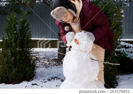 Full length portrait woman have fun, building snowman in snow covered backyard, putting carrot and tree branch on a huge snowball, enjoying happy winter weekend on snowy cool february day. Wintertime 103576931