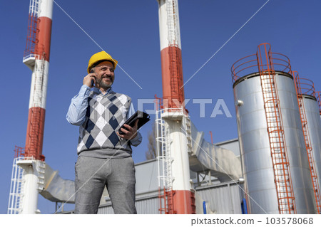 Smiling Businessperson in Yellow Hardhat Talking on Mobile Phone and Standing Against Power Plant Chimneys and Blue Sky Background Smiling Businessperson in Yellow Hardhat Talking on Mobile Phone and Standing Against Power Plant Chimneys and Blue Sky Background 103578068