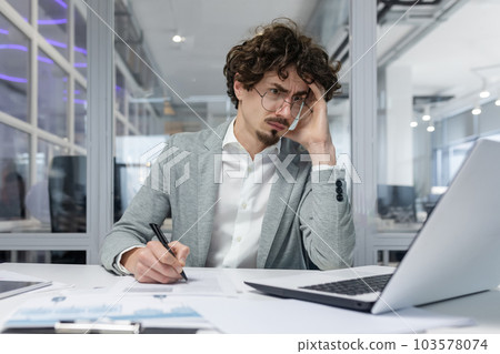 Upset businessman behind paper work inside modern office, mature man with beard reading financial reports and account documents unhappy with results and disappointed with achievements 103578074