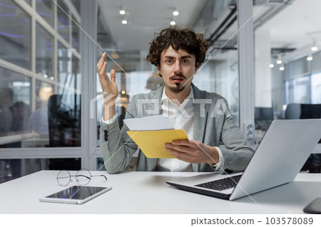 Upset businessman at workplace received letter with bad news, man looking at camera thinking and depressed, worker inside office with laptop. 103578089