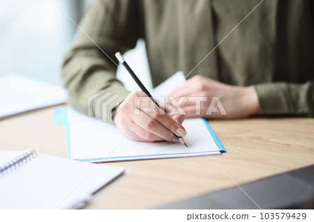 Woman writes on paper sheet with pencil sitting at desk 103579429