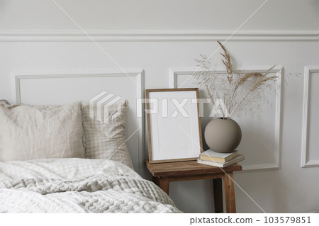 Elegant boho bedroom interior. Vertical wooden picture frame mockup, vintage bedside table. Ball vase with dry grass on old books. Beige linen throw bedding. White wall background. Scandinavian home. 103579851
