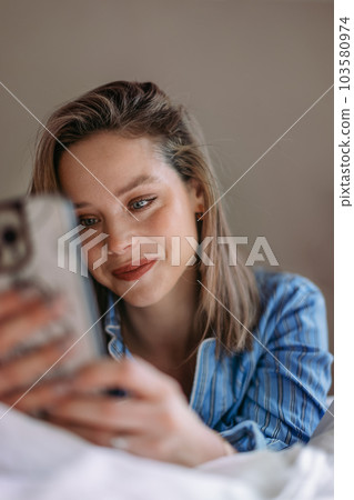 Young woman lying on a bed and scrolling her smartphone. Young woman lying on a bed and scrolling her smartphone. 103580974