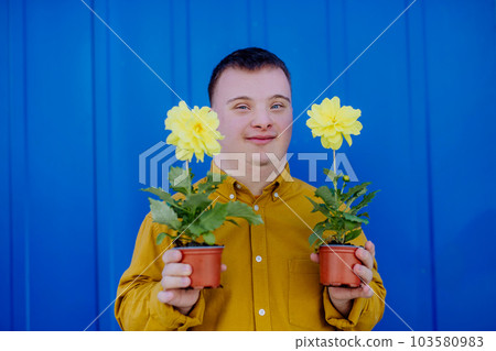 Happy young man with Down syndrome looking at camera and holding pot flowers against blue background. 103580983