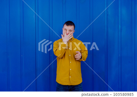 Happy young man with Down syndrome smiling and looking at camera against blue background. 103580985