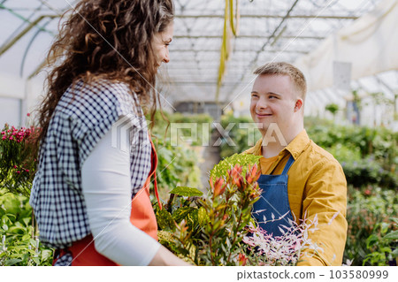 Experienced woman florist helping young employee with Down syndrome in garden centre. Experienced woman florist helping young employee with Down syndrome in garden centre. 103580999