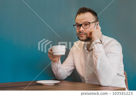Stylish young guy businessman from Europe drinks coffee and talks on his smartphone sitting at a table in a cafe on a break Stylish young guy businessman from Europe drinks coffee and talks on his smartphone sitting at a table in a cafe on a break 103581223