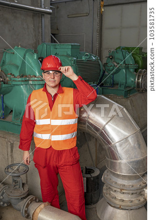 Young Technician in Red Coveralls and Hardhat Standing in Industrial Interior With Pipes and Generators Young Technician in Red Coveralls and Hardhat Standing in Industrial Interior With Pipes and Generators 103581735