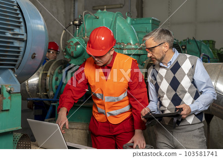 Young Technician with Laptop Computer Checking Data With Technical Manager in Mechanical Room 103581741