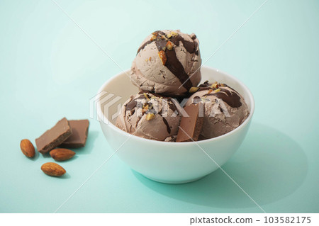Chocolate ice cream decorated with nuts and chocolate icing, in a white bowl, blue background. Summer homemade cooling desserts Chocolate ice cream decorated with nuts and chocolate icing, in a white bowl, blue background. Summer homemade cooling desserts 103582175
