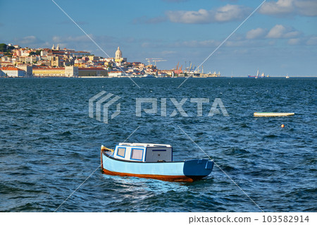 View of Lisbon over Tagus river from Almada with blue fishing boat and yachts tourist boats in background at sunset. Lisbon, Portugal View of Lisbon over Tagus river from Almada with blue fishing boat and yachts tourist boats in background at sunset. Lisbon, Portugal 103582914