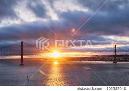 View of 25 de Abril Bridge famous tourist landmark of Lisbon connecting Lisboa and Almada on Setubal Peninsula over Tagus river with tourist yacht boats at sunset. Lisbon, Portugal 103582948