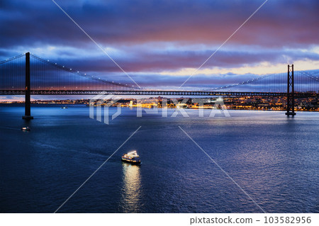 View of 25 de Abril Bridge famous tourist landmark of Lisbon connecting Lisboa and Almada on Setubal Peninsula over Tagus river in the evening twilight with tourist boats. Lisbon, Portugal 103582956