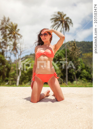 Young woman on her knees in beach sand, shading her eyes from sun, green palms jungle in background 103583474