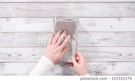 Step by step. Folding natural color linen dinner napkin on a wooden white table. Step by step. Folding natural color linen dinner napkin on a wooden white table. 103583579