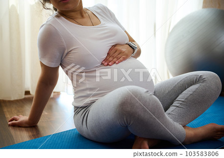 Close-up pregnant millennial woman sitting on exercise mat, touches her belly after performing prenatal and meditation exercises at cozy tranquil homely atmosphere. Pregnancy and maternity lifestyle 103583806