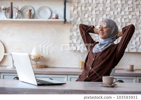 Rest after distance learning. Young Muslim female student in hijab sits at home at the table in front of the laptop and relaxes. She threw her hands behind her head, closed her eyes, a break. Rest after distance learning. Young Muslim female student in hijab sits at home at the table in front of the laptop and relaxes. She threw her hands behind her head, closed her eyes, a break. 103583931