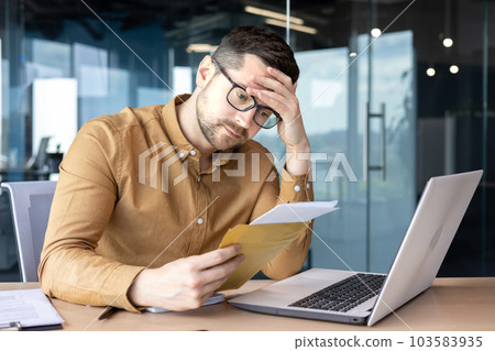 Shocked young man businessman sits in the office at the table and reads the received letter, documents. He holds his head worriedly. Shocked young man businessman sits in the office at the table and reads the received letter, documents. He holds his head worriedly. 103583935