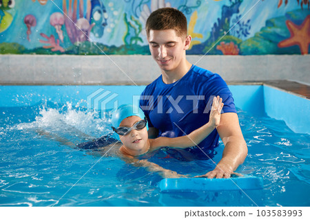 Trainer teaching little boy how to swim in indoor pool with pool floating board 103583993