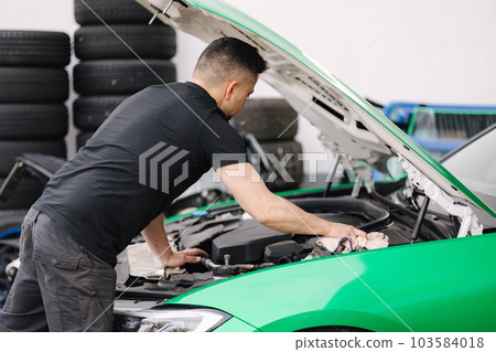 Portrait of a male mechanic working in his garage. Young bearded man checks the engine in the car, the hood is open Portrait of a male mechanic working in his garage. Young bearded man checks the engine in the car, the hood is open 103584018