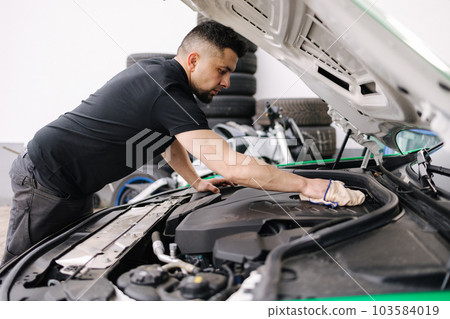 Side view of a male mechanic working in his garage. Young bearded man checks the engine in the car, the hood is open 103584019