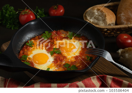 Shakshuka from two eggs in tomato sauce with fresh tomatoes, spices and herbs in a black frying pan. Close-up scrambled eggs 103586404