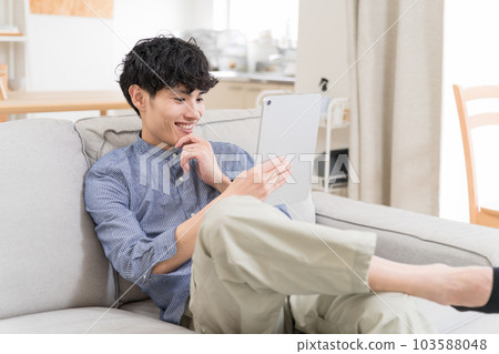 年輕人坐在沙發上看著平板電腦笑著 103588048