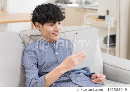 年輕人坐在沙發上看著平板電腦笑著 103588049