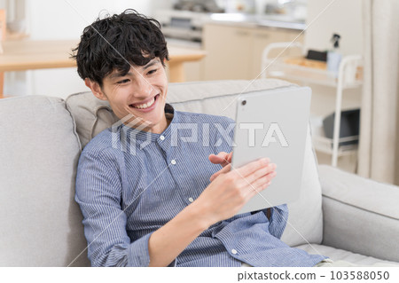 年輕人坐在沙發上看著平板電腦笑著 103588050