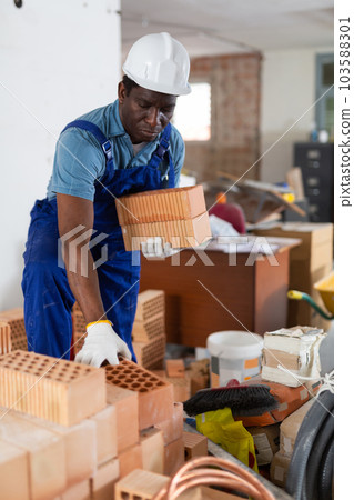 Builder in overall and hardhat carrying bricks in apartment 103588301