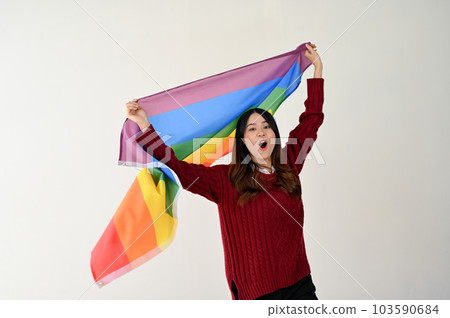 A cheerful Asian woman with a rainbow LGBT flag stands over an isolated white background. 103590684