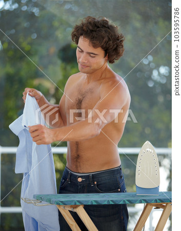Iron, shirt and a topless man doing laundry outdoor on the deck of his home for housework during the day. Clothes and ironing board with a handsome young male person doing household chores 103595786