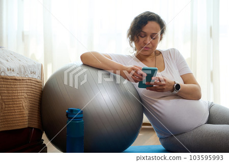 Young pregnant woman wearing smart watch to control pulse and heart rate, relaxing with smartphone on yoga mat after training at home, browsing mobile application, resting after meditation practice Young pregnant woman wearing smart watch to control pulse and heart rate, relaxing with smartphone on yoga mat after training at home, browsing mobile application, resting after meditation practice 103595993