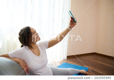 Beautiful adult pregnant woman having rest after prenatal fitness at home, holding mobile phone, making a self-portrait, sharing happy moments of her active healthy pregnancy lifestyle in social media Beautiful adult pregnant woman having rest after prenatal fitness at home, holding mobile phone, making a self-portrait, sharing happy moments of her active healthy pregnancy lifestyle in social media 103595995