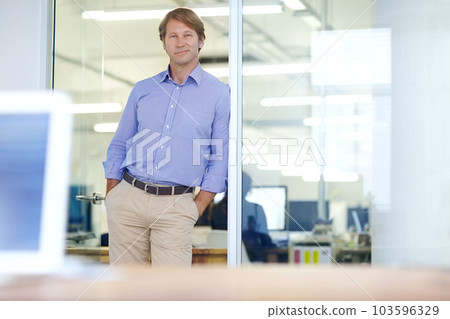 Surveying his workplace. Mature businessman leaning on an office doorway and smiling slightly with his hands in his pockets. 103596329