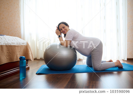 Happy adult pregnant woman leading active healthy lifestyle in pregnancy time, doing stretching exercises, standing on knees on a yoga mat leaning on a fitness ball at home, smiling looking at camera 103598096