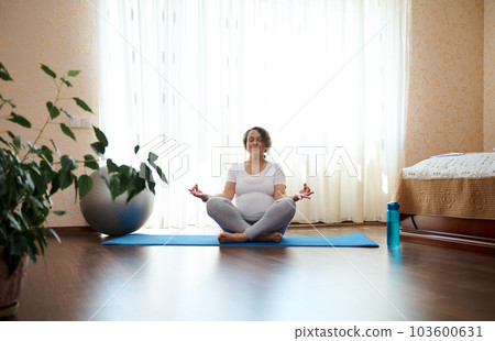 Authentic relaxed pregnant woman meditating in lotus pose with mudra gesture on yoga mat, in cozy home bedroom, against large window on sunny day. Healthy active lifestyle, wellness in pregnancy time 103600631