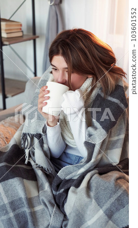 Fever disease. Sick woman. Home recovery. Shivering lady wrapped in plaid sitting sofa drinking tea in light room interior . 103601552