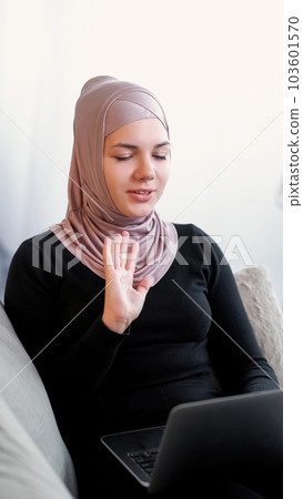 Video call. Online conference. Internet communication. Relaxed young woman in hijab and black clothes talking to laptop waving hand greeting in light room at home. Video call. Online conference. Internet communication. Relaxed young woman in hijab and black clothes talking to laptop waving hand greeting in light room at home. 103601570