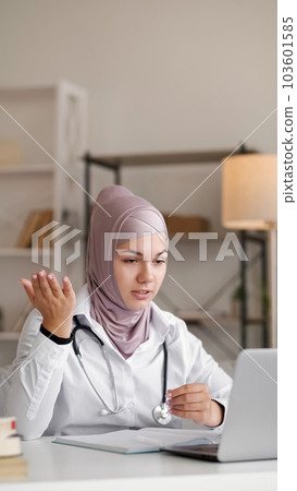 Online doctor. Video connection. Virtual healthcare. Female medical worker woman in hijab with stethoscope talking at conference gesticulating at table with laptop in white room. Online doctor. Video connection. Virtual healthcare. Female medical worker woman in hijab with stethoscope talking at conference gesticulating at table with laptop in white room. 103601585