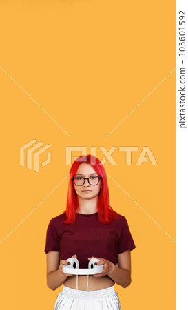 Music device. Modern gadget. Young ginger girl in glasses with beautiful contemporary design wire headphones in hands on orange background empty space. Music device. Modern gadget. Young ginger girl in glasses with beautiful contemporary design wire headphones in hands on orange background empty space. 103601592