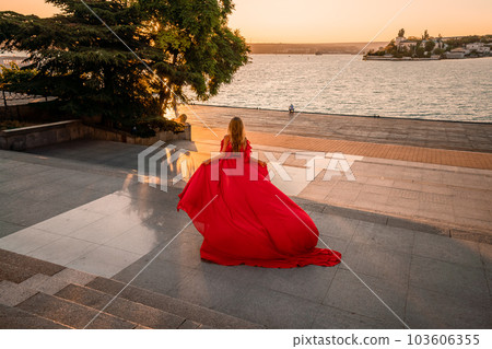 Sunrise red dress. A woman in a long red dress against the backdrop of sunrise, bright golden light of the sun's rays. The concept of femininity, harmony. 103606355