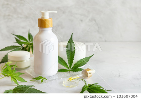 White One pump bottle near green cannabis leaves on white table. Cosmetic Mockup White One pump bottle near green cannabis leaves on white table. Cosmetic Mockup 103607004