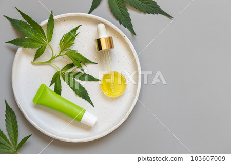 Cream tube and pipette near green cannabis leaves on grey table. Cosmetic Mockup 103607009