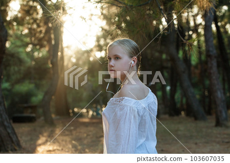 Pensive teenage girl in white shirt listen to music in headphones on the background of forest 103607035