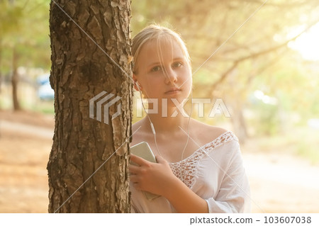 Teenage girl in headphones holding mobile phone and standing near tree on the background of sea 103607038